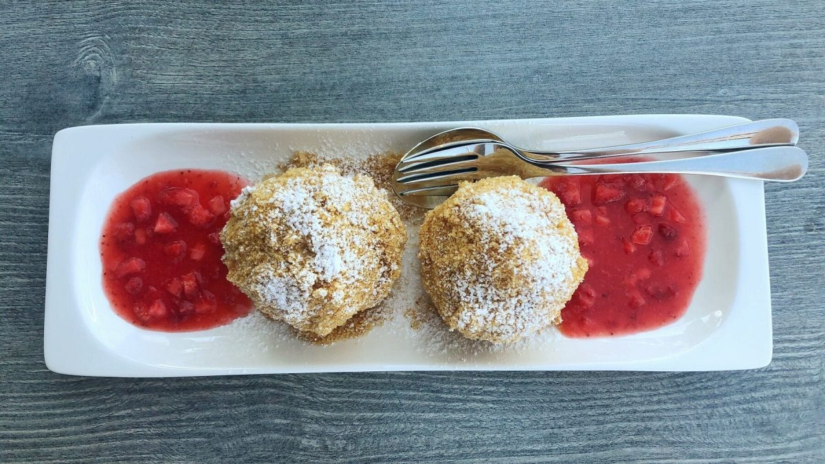Zwei Topfenknödel mit Erdbeer-Rhabarber-Kompott auf einem länglich-weißen Teller mit Besteck