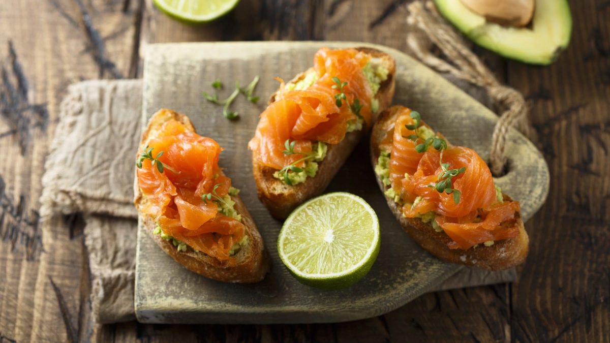 Avocado-Lachs-Bruschetta, garniert mit Limettensaft und frischer Kresse, serviert auf einem Holzbrett auf einem dunklen Holztisch.