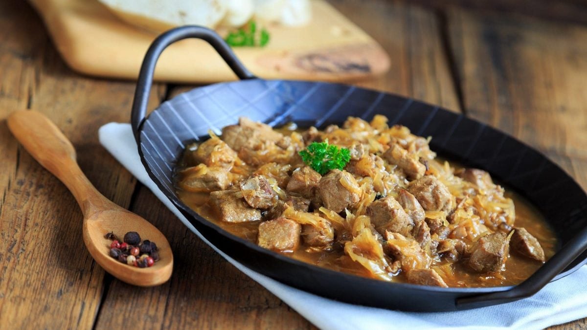 In einer rustikalen Pfanne wird original Szegediner Gulasch serviert. Neben der Pfanne liegen Pfefferkörner . Die Pfanne steht auf einem rustikalem Tisch.
