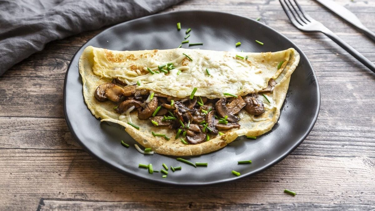 Deftige Pfannkuchen mit Champignons und Käsestreuseln gefüllt, serviert auf einem schwarzen Teller auf einem grauen Holztisch.
