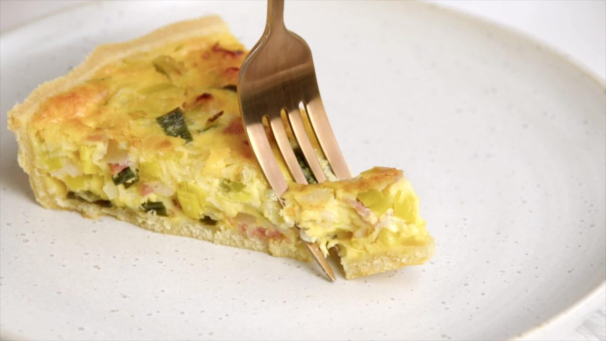 Auf einem Teller ist ein Stück Quiches Lorraine zu sehen. Eine Gabel sticht in das Stück.