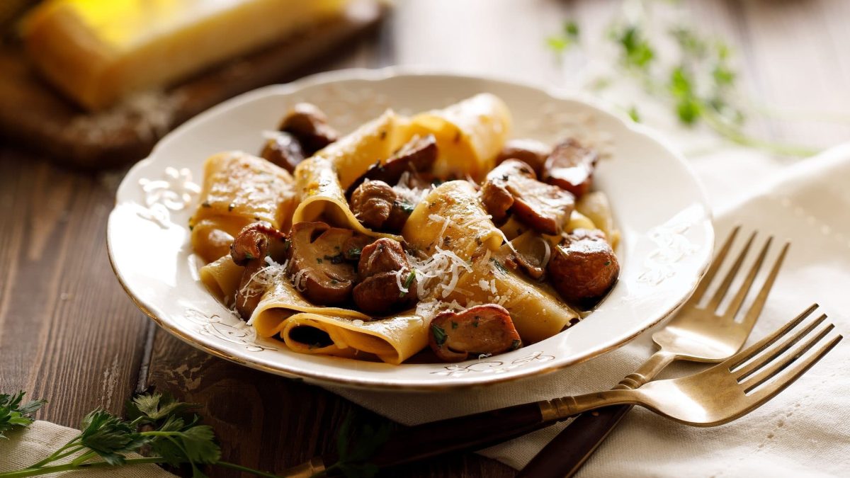 Auf einem rustikalem Holztisch steht ein weißer Teller mit Pappardelle mit Kraeutern und Pilzen.