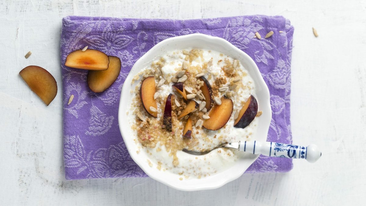 Pflaumen-Frühstücksbrei aus Quinoa mit Joghurt in weißer Schüssel auf lila Tuch