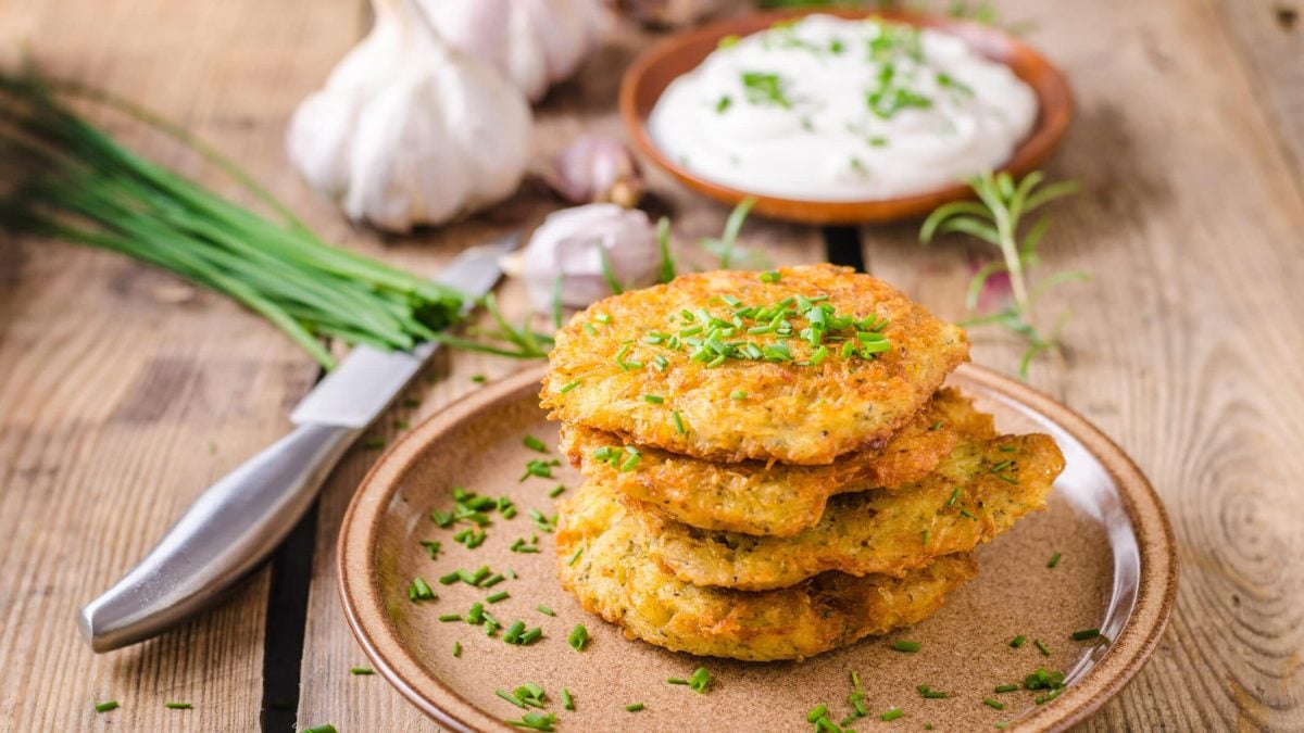 Kartoffelpuffer mit selbstgemachtem Quarkdip auf braunem Teller mit Schnittlauch, dahinter Knoblauch und Messer