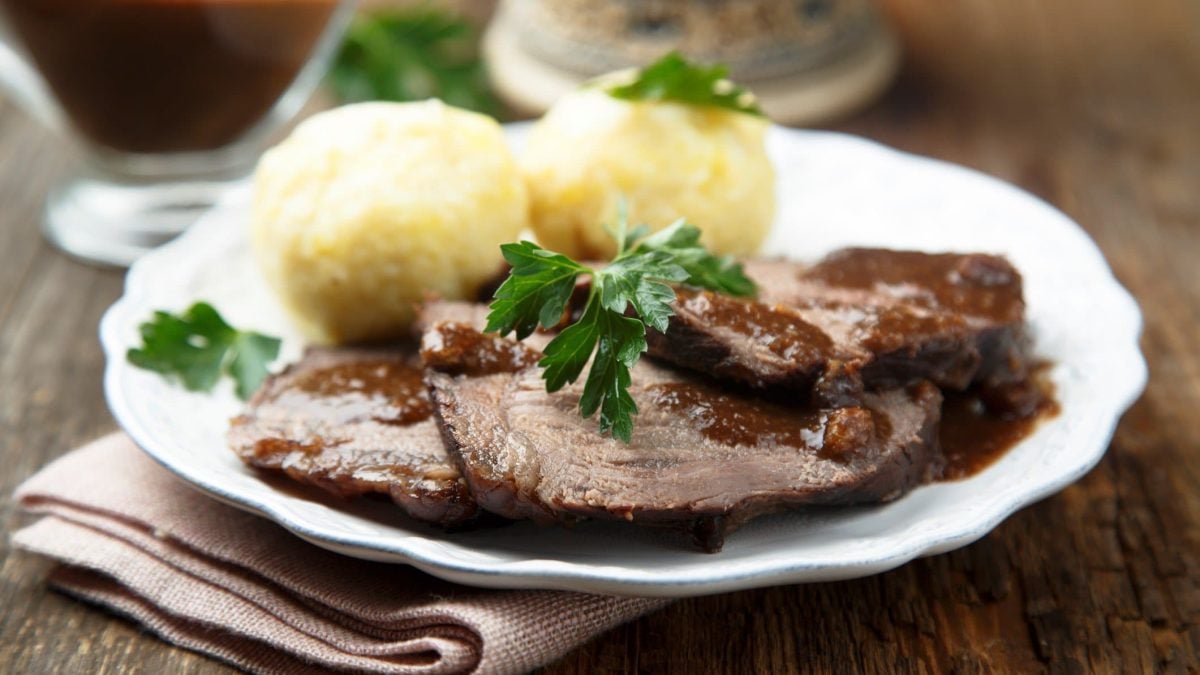 Sauerbraten aus Kalbsbäckchen mit Soße und Klößen auf weißem Teller, beigem Tuch und dunklem Tisch. Garniert mit Petersilie. Im Hintergrund Sauciere aus Glas mit Soße. Aufnahme aus der Frontalsicht.