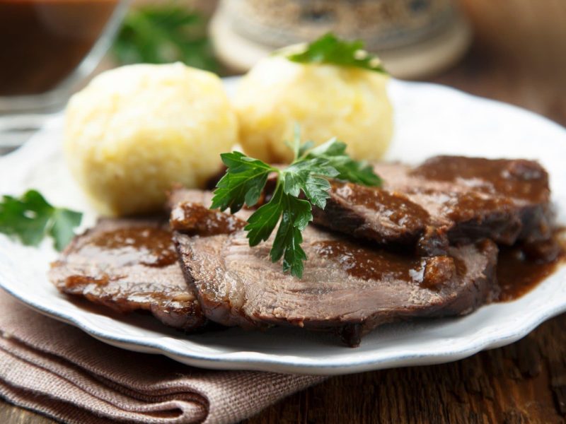 Sauerbraten aus Kalbsbäckchen mit Soße und Klößen auf weißem Teller, beigem Tuch und dunklem Tisch. Garniert mit Petersilie. Im Hintergrund Sauciere aus Glas mit Soße. Aufnahme aus der Frontalsicht.
