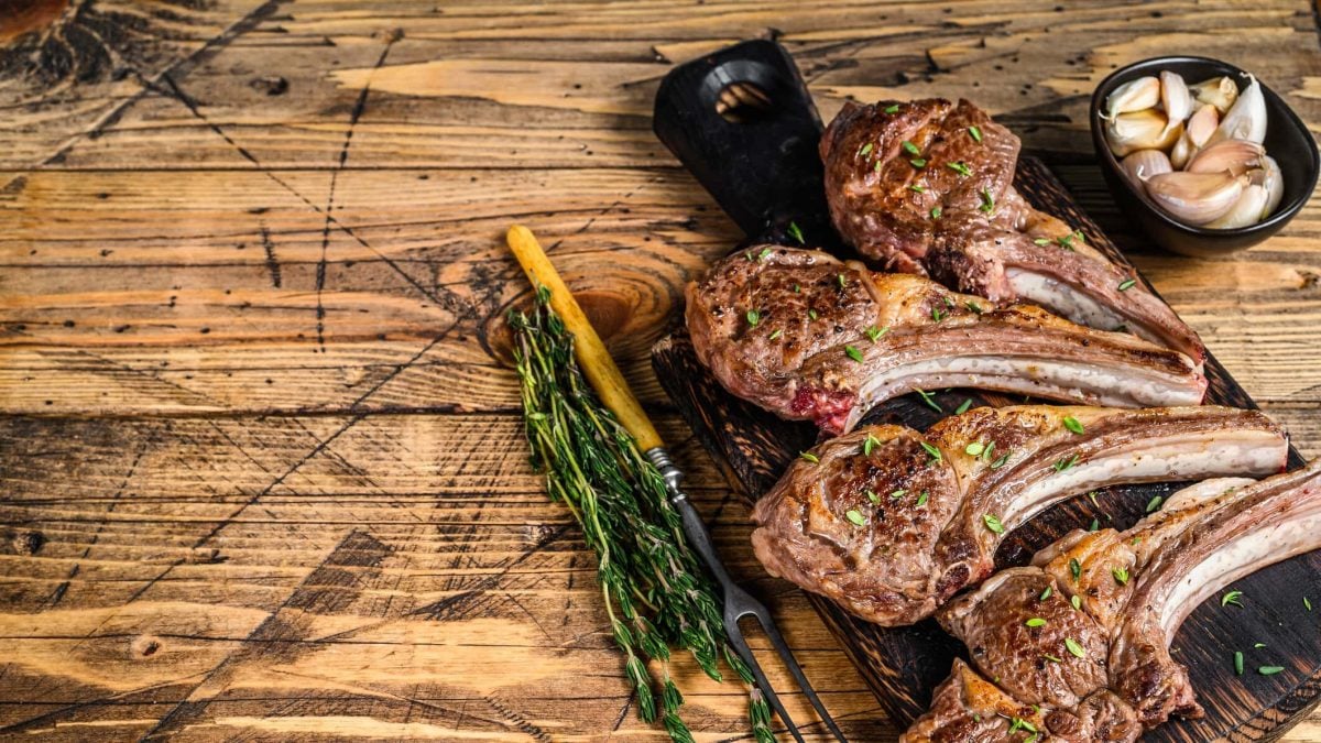 vier gegrillte Lammkoteletts auf Brett, garniert mit einem Stängel Rosmarin und Knoblauchzehen im Hintergrund