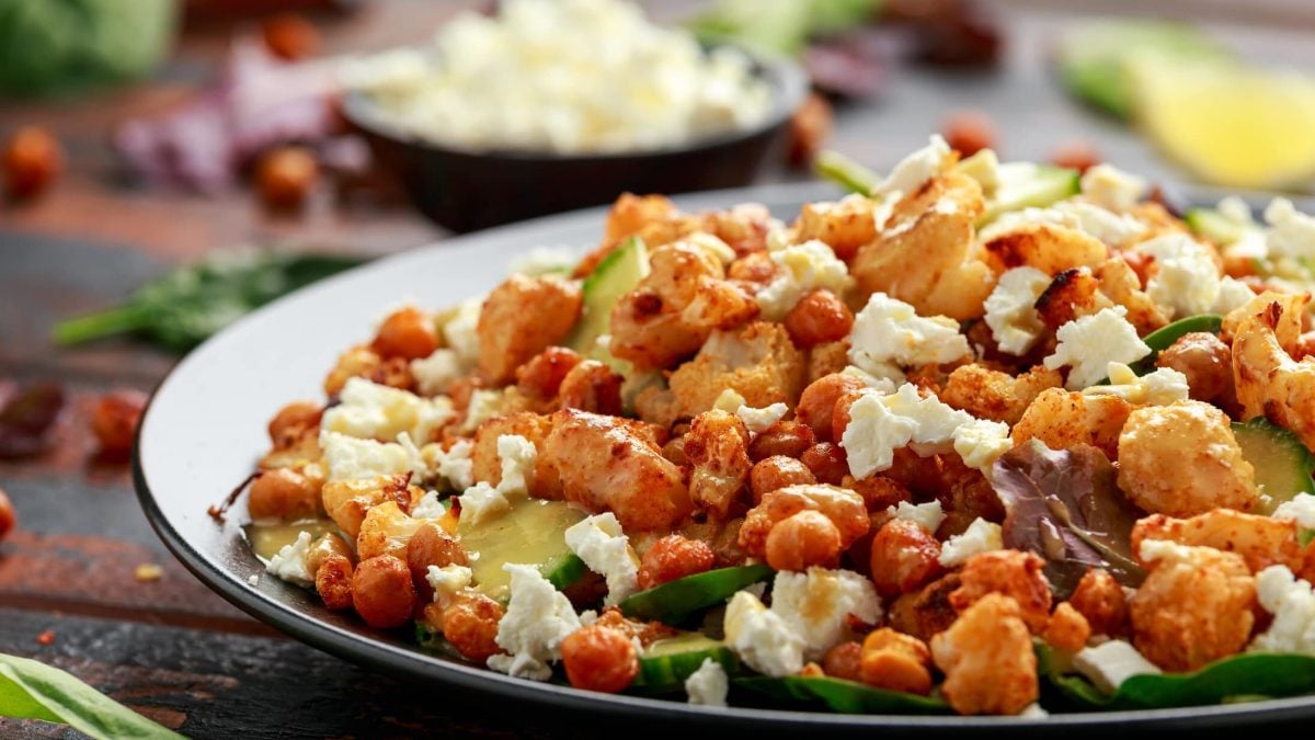 Salat mit geröstetem Blumenkohl, Gurke und Kichererbsen auf dunkler Platte, darüber Feta gebröselt