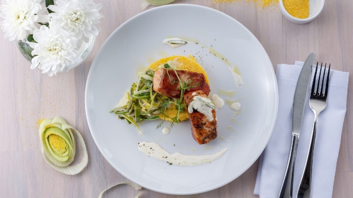 Gegrilltes Lachsfilet auf Polenta-Talern auf einem weißen Teller neben Besteck und Porree
