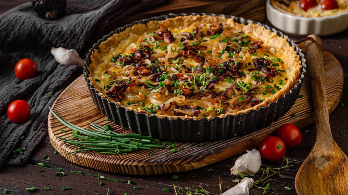 Vegetarischer Zwiebelkuchen mit Räuchertofu auf Holzuntergrund, daneben tomaten und Lräuter