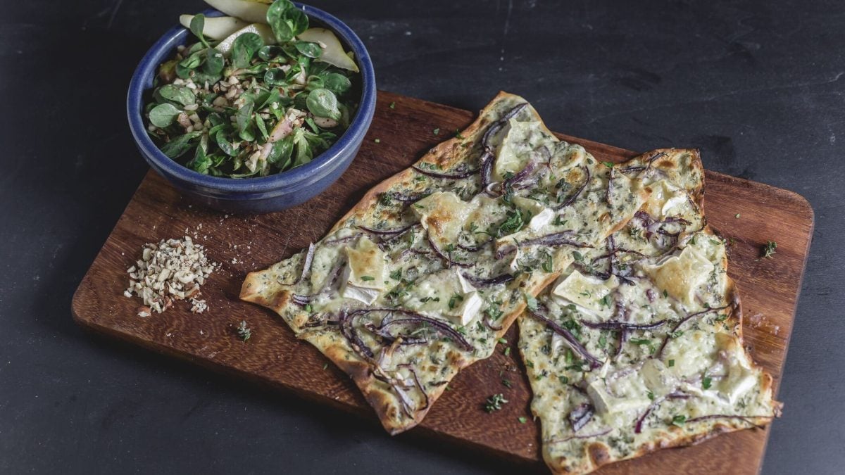 Flammkuchen mit Birne und Ziegenkäse auf Holzbrett, dahinter Salatschüssel