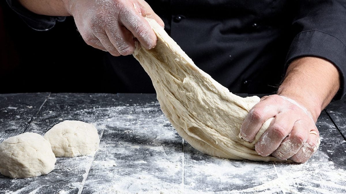 Auf einer bemehlzen Arbeitsfläche kneten ein Mann einen Pizzateig. Von dem Mann sind nur die Hände zu sehen.