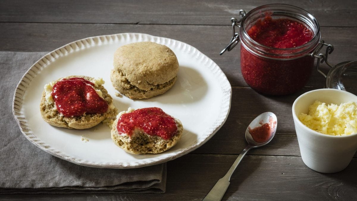 Britische Scones mit Erdbeermarmelade vor Holzuntergrund auf weißem Teller, daneben Marmelade und Clotted Cream