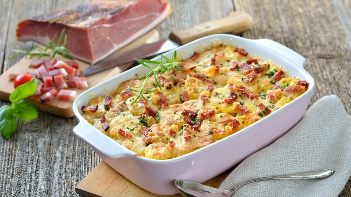 Draufsicht: Hölzerner Untergrund. Im Hintergrund ein Stück frischer Speck, teilweise in Würfel geschnitten. Im Bildvordergrund eine Auflaufform mit Kartoffel-Kohlrabi-Gratin mit Speck.