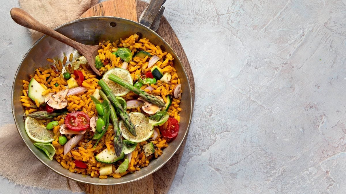 Bunte vegane Gemüse-Paella mit verschiedenen Gemüsesorten, garniert mit Zitronenhälften und Oregano in einer großen Kupferschale auf hellem Untergrund
