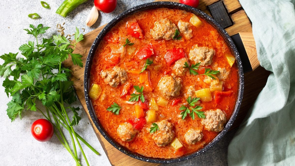 Linsen-Tomatensuppe mit Fleischbällchen, Paprika und frischer Petersilie, in einem großen Topf auf hellem Holzbrett serviert.