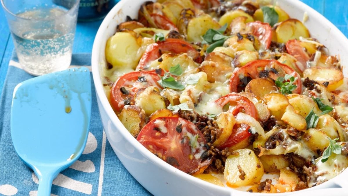 Schrägsicht: Blauer Untergrund mit einer ovalen Auflaufform mittig-rechts im Bild. Gefüllt mit einem Bratkartoffel-Hack-Auflauf mit frischen Tomaten. Links daneben ein blauer Pfannenwender.