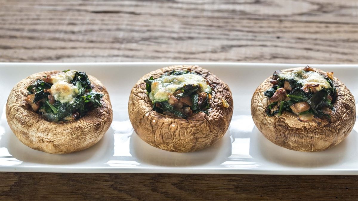 Mit Käse überbackene gefüllte Champignons mit Spinat, Speck und Zwiebeln, nebeneinander audgereiht auf einem weißen Teller auf einem hölzernen Tisch.