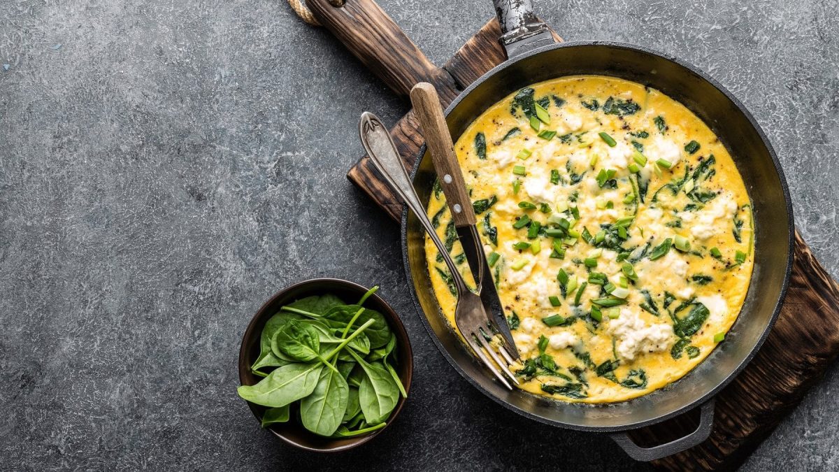 Aus der Vogelperspektive ist eine Pfanne zu sehen. IN der Pfanne ist ein Spinat-Feta-Omelett. Daneben liegen Messer und Gabel, sowie ein Schöächen mit Spinatblättern.