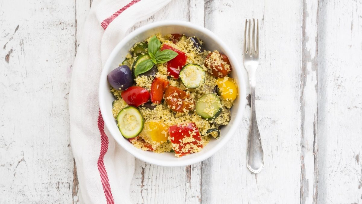 Couscous-Zucchini-Pfanne mit Oliven, gebratenen Zwiebeln, Paprika und frischem Basilikum in einer Schale auf einem hellen Holztisch serviert.