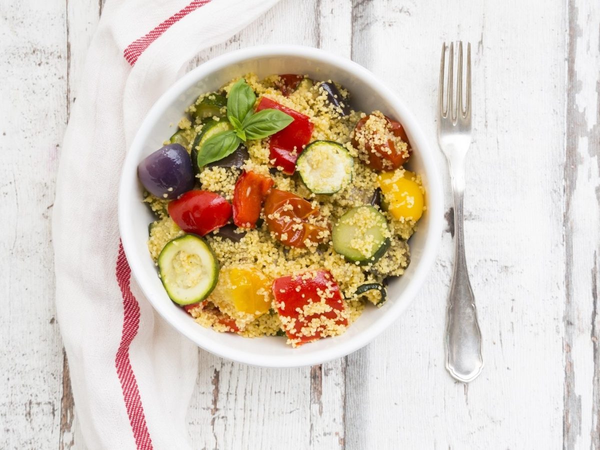 Couscous-Zucchini-Pfanne mit Oliven, gebratenen Zwiebeln, Paprika und frischem Basilikum in einer Schale auf einem hellen Holztisch serviert.