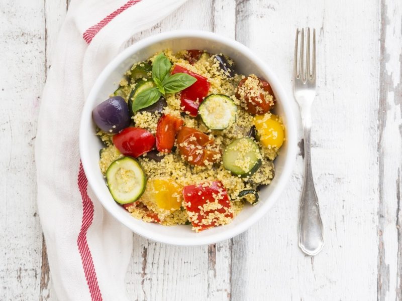 Couscous-Zucchini-Pfanne mit Oliven, gebratenen Zwiebeln, Paprika und frischem Basilikum in einer Schale auf einem hellen Holztisch serviert.