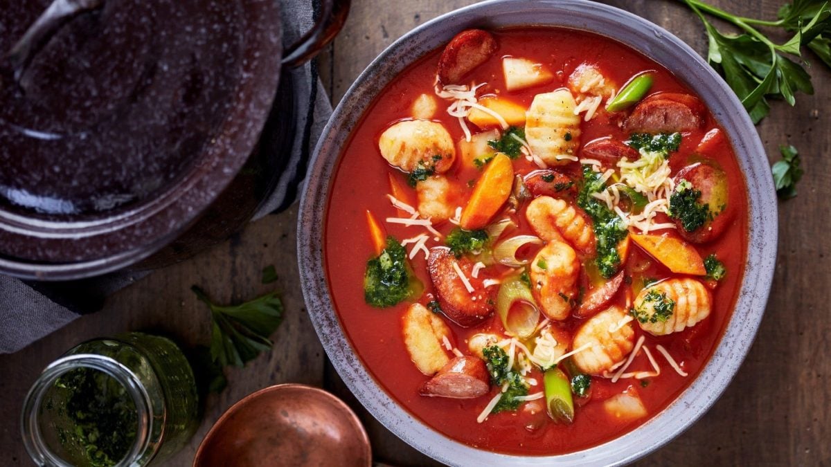 One-Pot-Gnocchi mit Wurst, Gemüse und Kräutern in einer deftigen Tomatensuppe als Eintopf, serviert in einer Tonschale auf hölzernem Untergrund
