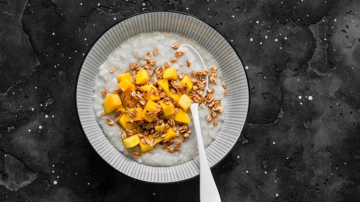 Haferbrei mit Kokos und Mangostückchen in einer Bowl, bestreut mit angerösteten Haferflocken und serviert auf einem dunklen Schiefertisch.