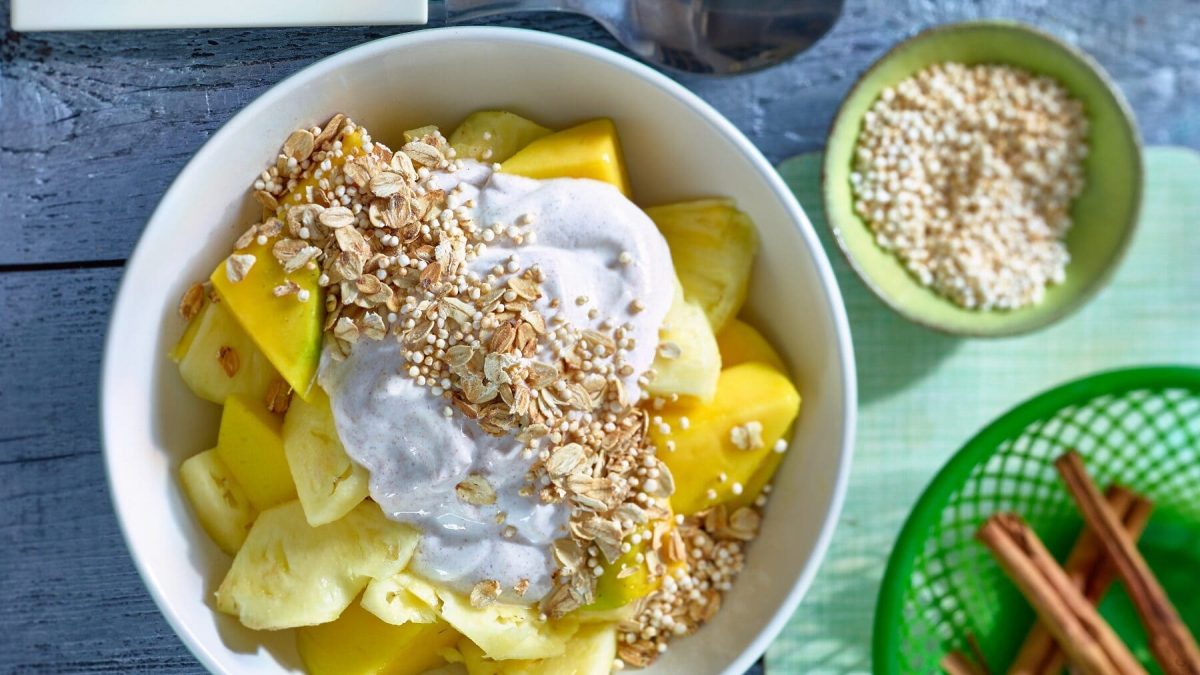 Eine Schale mit Ananas-Mango-Granola neben kleinen Schälchen mit Zutaten auf hellblauem Hintergrund.