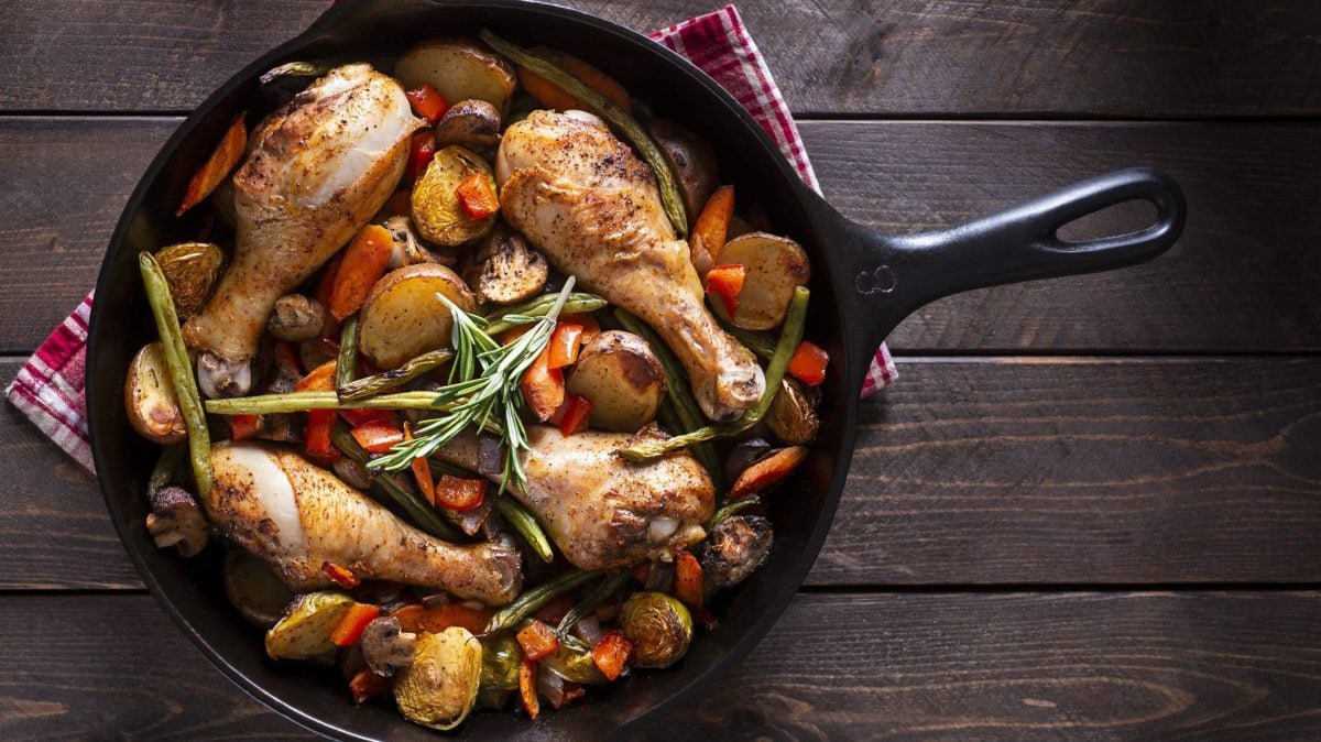 Rustikale Wok-Pfanne mit Hähnchenkeulen und Rosmaringemüse auf dunklem Holzuntergrund