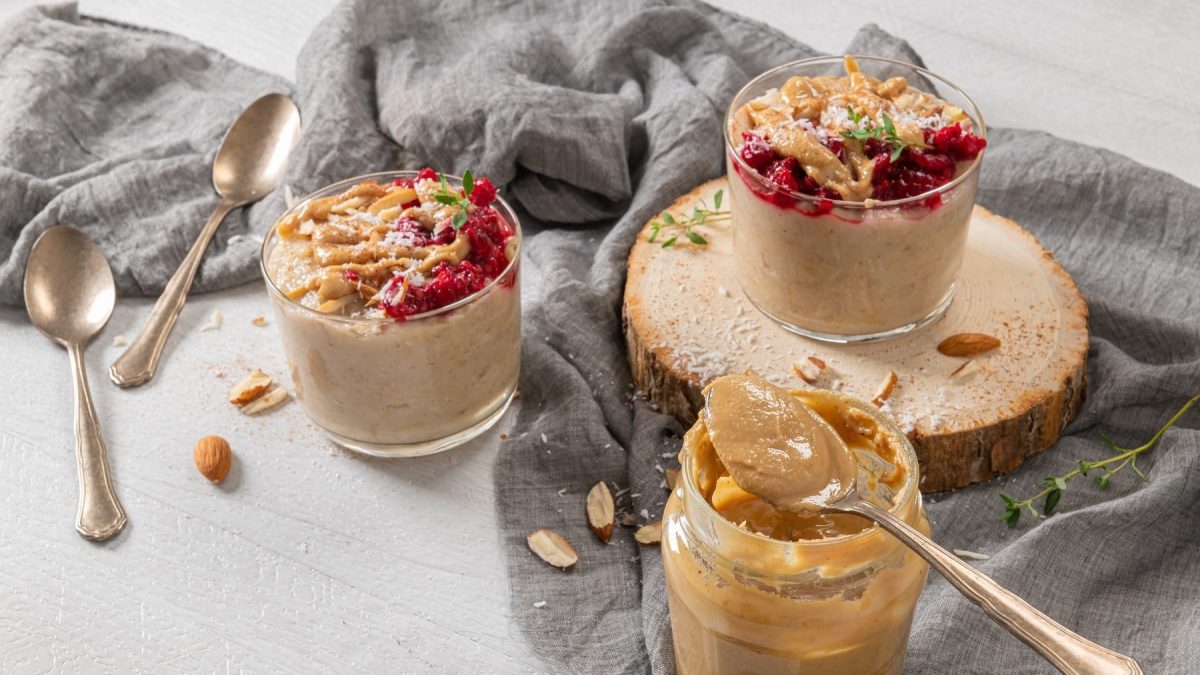 Porridge mit Himbeeren und Mandelmus, Kokosflocken und frischem Oregano garniert, serviert in Dessertschälchen auf einem grauen Tuch.