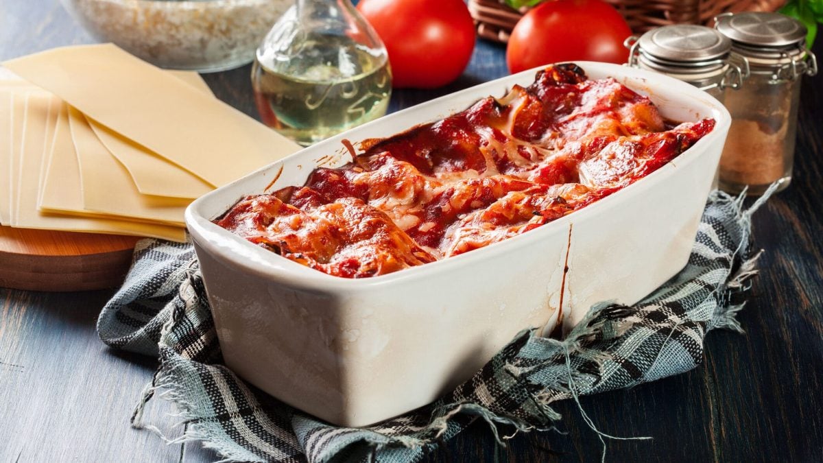 Weiße Kastenform mit Bacon-Lasagne, dahinter Lasagneblätter und Zutaten