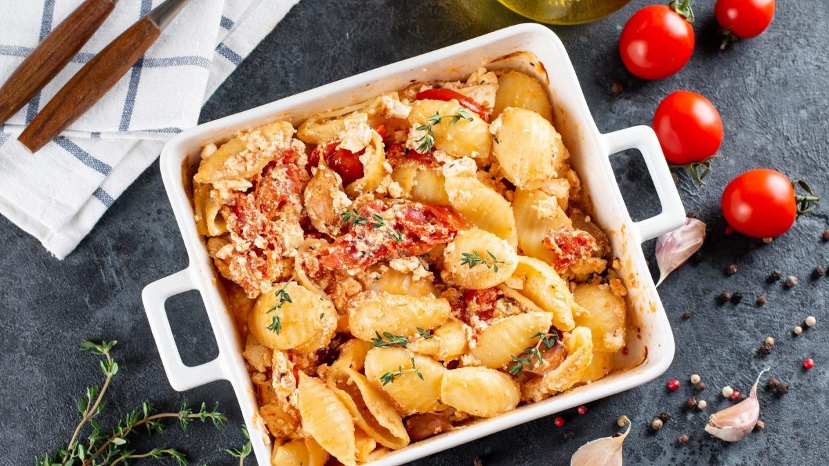 Baked Feta Pasta in einer weißten Auflaufform mit frischem Thymian auf einem graunen Schiefertisch mit frischem Pfeffer, Knoblauch und Tomaten als Deko.