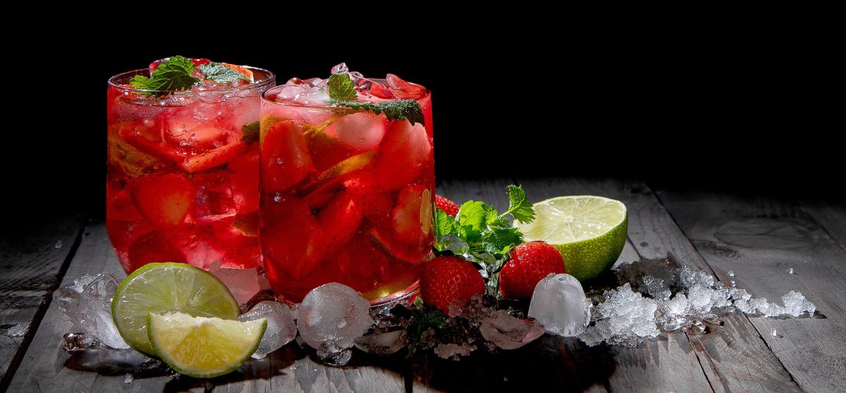 Erdbeer-Mojito in zwei Gläsern auf dunklem Untergrund, daneben Crushed Ice, Erdbeeren, Minze und Limetten.