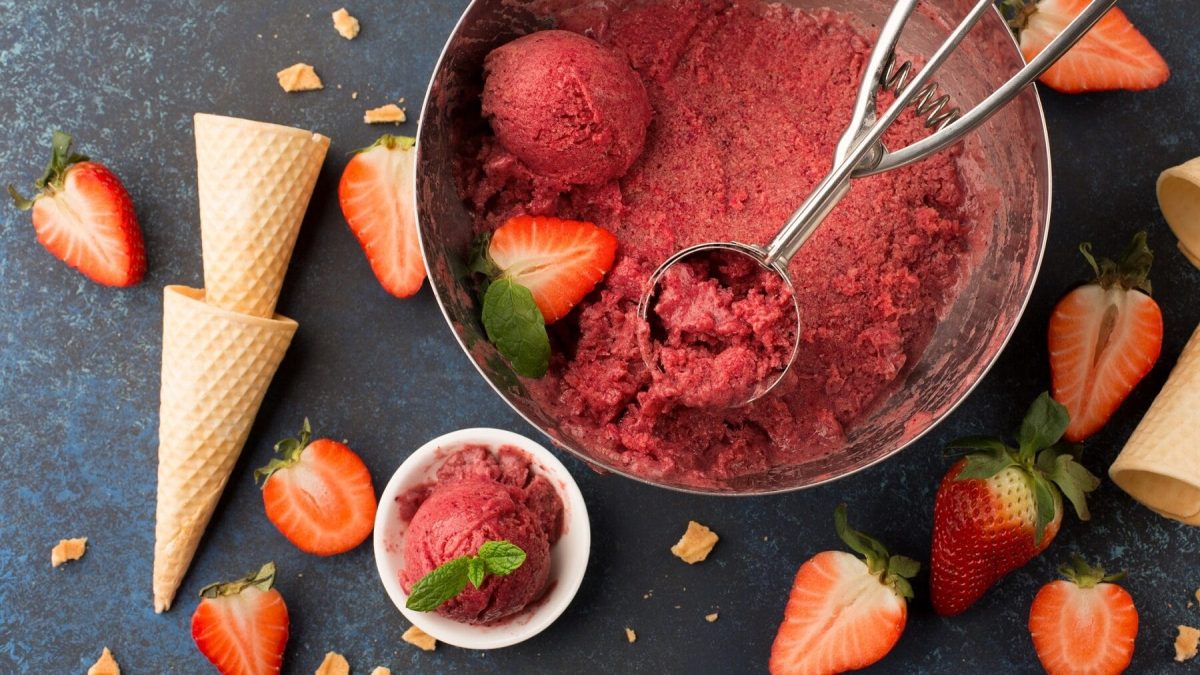 Eine große Metallschüssel mit Erdbeersorbet steht auf einem blau-grauen Untergrund. Neben der Metallschüssel steht eine kleinere Porzellanschüssel mit einer Portion Sorbet. In der Metallschüssel befindet sich ein Eisportionierer. Auf dem Untergrund liegen Eiswaffeln und Erdbeeren.