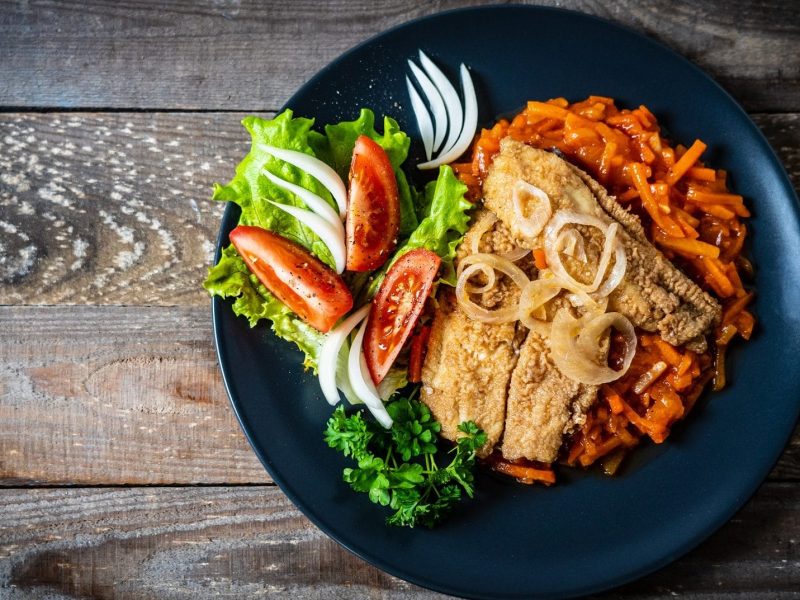 Gebratenes Fischfilet mit Tomatensauce : ein Teller mit Tomaten-Möhren-Soße, Fisch und Salat auf einem Holztisch.