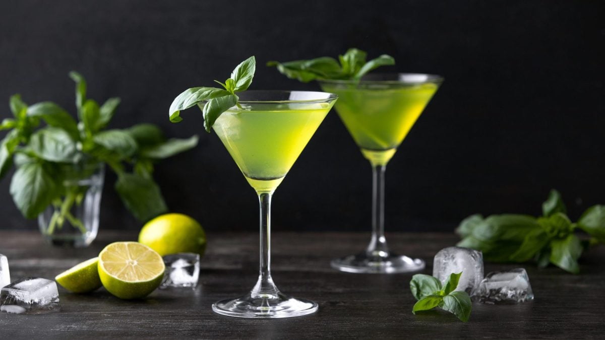 Zwei Gläser mit Gin-Basil-Smash stehen auf einem dunklen Untergrund vor einer schwarzen Wand. Um die Gläser herum liegen Basilikumblätter, angeschnittene Zitronen und einige Eiswürfel. Die Cocktails sind mit Basilikumblättern angerichtet.