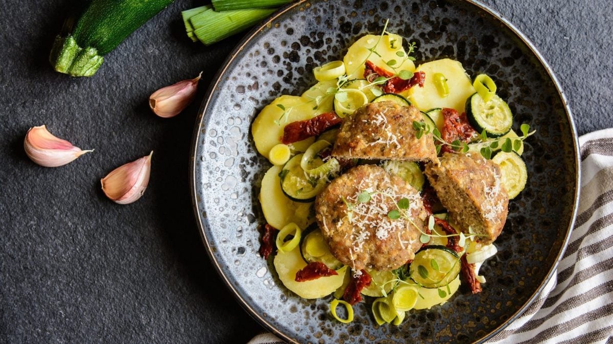 Griechische Hackbällchen auf einem Teller mit Zucchini und Kartoffelscheiben, dazu getrocknene Tomaten, Lauch und Kresse. Daneben Lauchstangen, eine Zucchini und Knoblauchzehen.