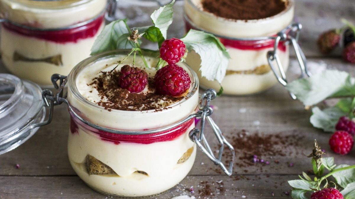 Mehrere verschließbare Gläser stehen auf einem grauen Tisch. Die Gläser sind gefüllt mit Himbeer-Tiramisu und bestreut mit Kakaopulver. Neben den Gläsern ist etwas Kakaopulver auf dem Tisch verstreut. Einige Himbeeren mit Blättern liegen dekorativ am Bildrand.