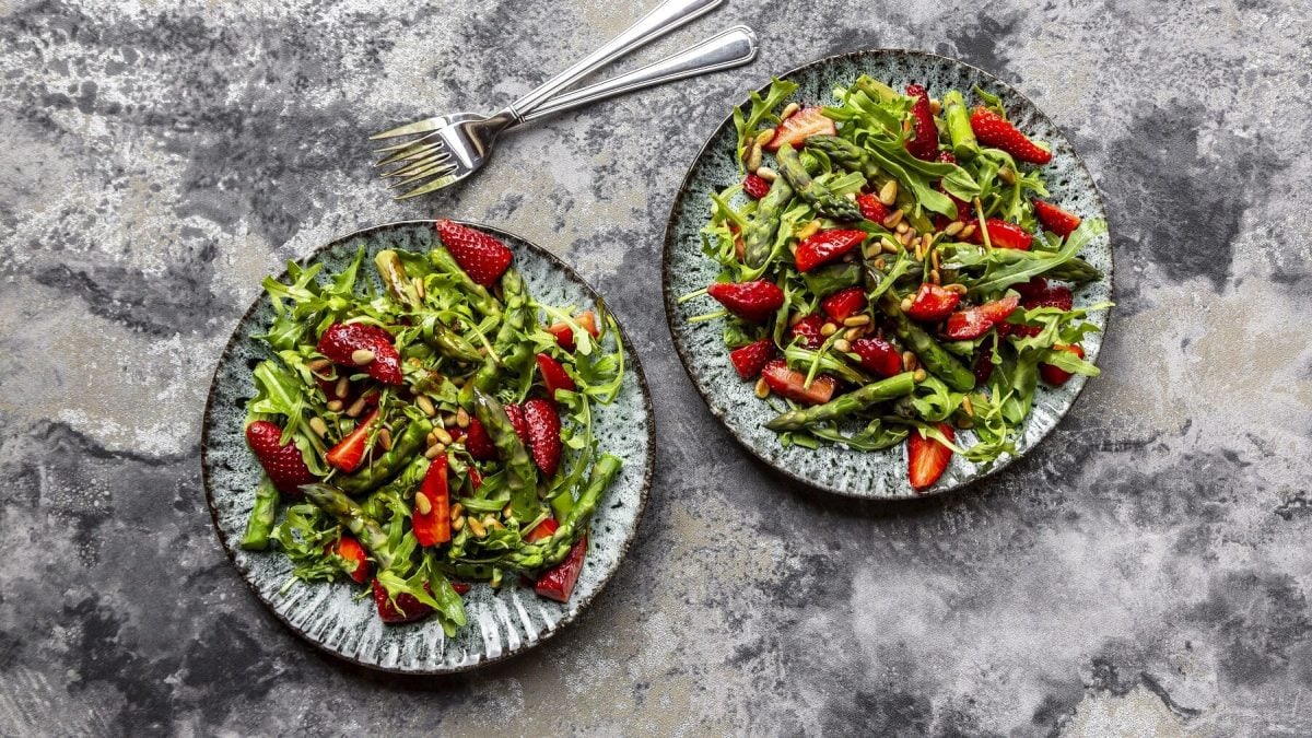 Bunter Spargelsalat mit Erdbeeren und Pinienkernen auf zwei Tellern auf einem grauen Steintisch neben zwei Gabeln.