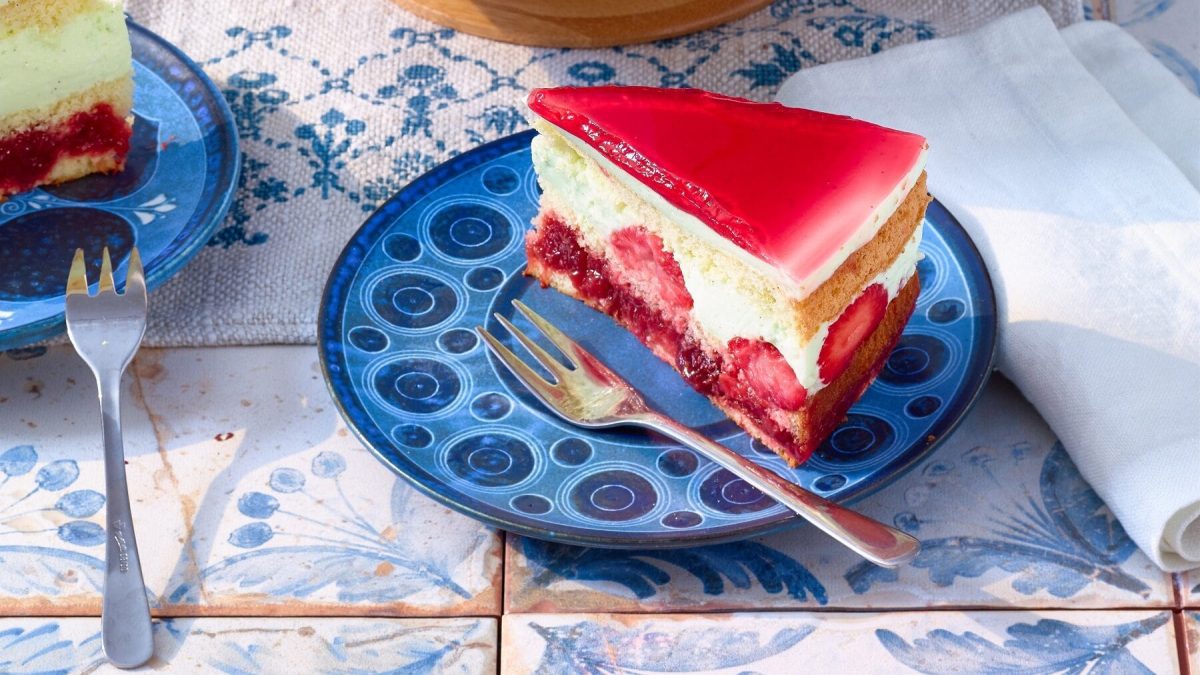 Sahnetorte mit Erdbeerbowle-Füllung mit frischen Erdbeeren und einem Erdbeerbowlen-Guss auf einem verzierten blauen Teller mit einer Gabel auf einem blauen Fliesentisch neben Papierservietten.