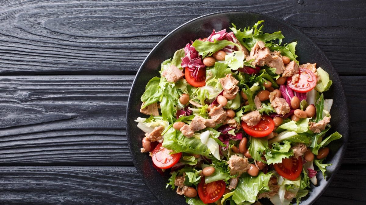 Eine Schüssel mit Thunfisch Tomaten Salat steht auf einem schwarzen Teller auf einem schwarzen Holztisch. Im Salat sind einige Bohnen verteilt.