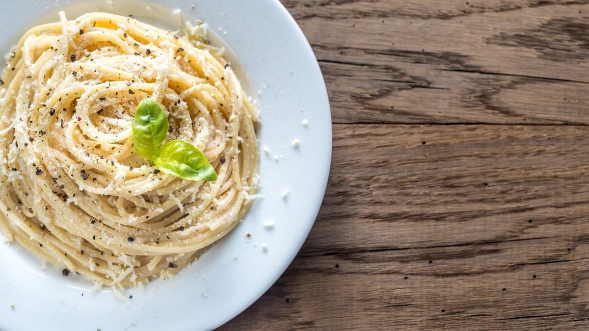 Pasta Cacio e Pepe in einem weißen Teller mit frischem Basilikum, geriebenem Parmesan und gemahlenem Pfeffer auf einem hellen Holztisch.