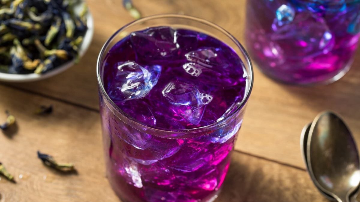 Ein Glas mit Schmetterlingsblüten Limonade mit Eiswürfeln auf einem hellen Holztisch mit frischen Schmetterlingsblüten und zwei Löffeln.