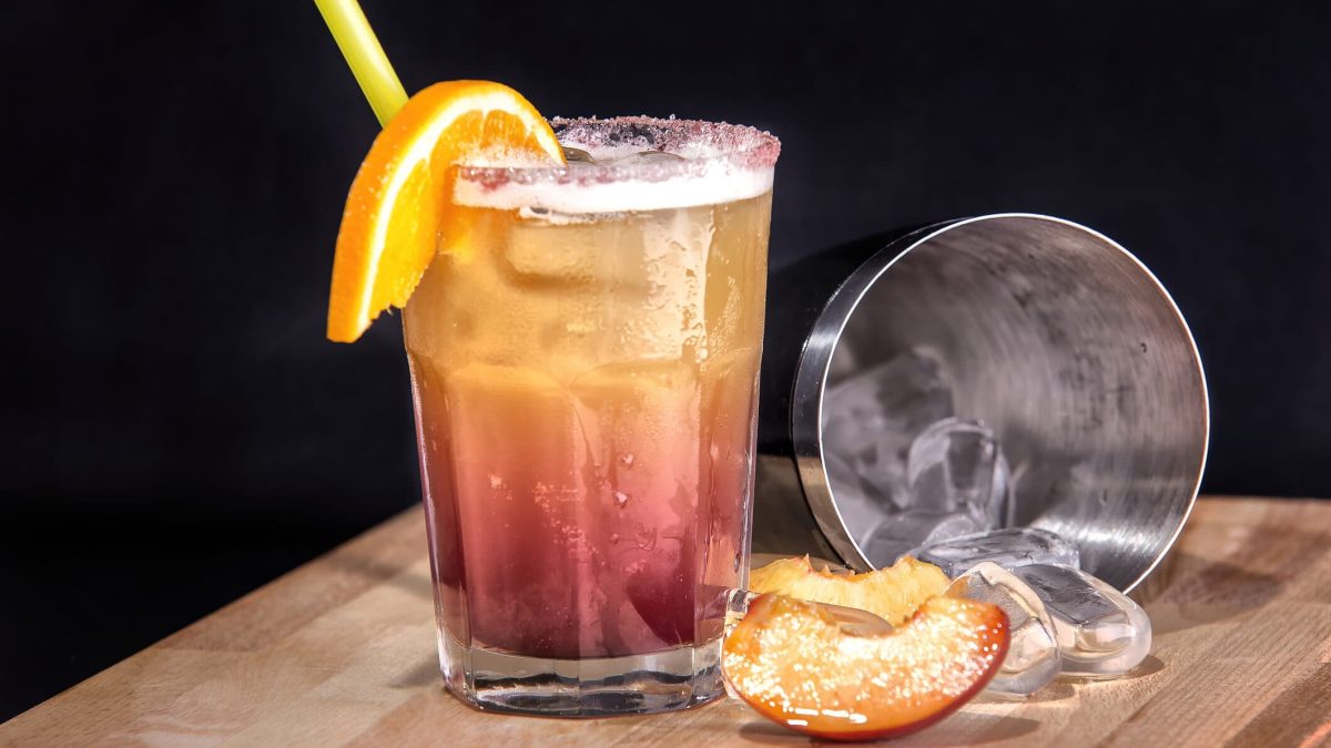 Ein Glas Sex on the Beach steht auf einem braunen Holzbrett vor einem schwarzen Hintergrund. Neben dem Glas liegt ein Mixgefäß mit Eiswürfeln und einige Pfirsichstücken. Das Glas ist dekoriert mit einer Orangenscheibe und ein gelber Stohhalm steckt im Glas.