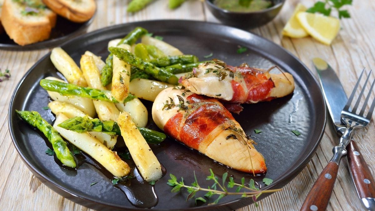 Gebratener grüner und weißer Spargel mit in Schinken gewickelter Hähnchenbrust auf einem schwarzen Teller neben Besteckt auf einem hellen Holztisch.