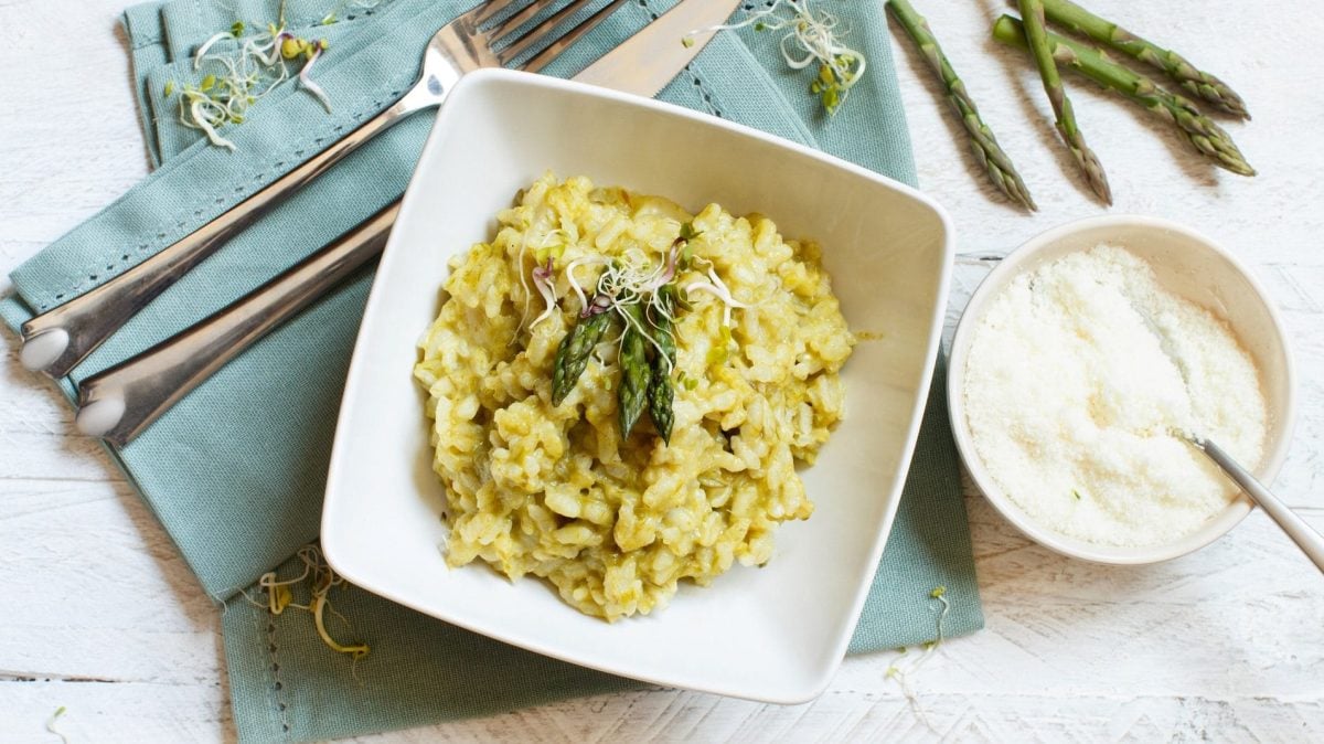 Risotto mit grünem Spargel: ein Teller mit Risotto und grünem Spargel auf einer Serviette, daneben eine Schüssel Parmesan, Besteck und ein paar Spargelstangen.