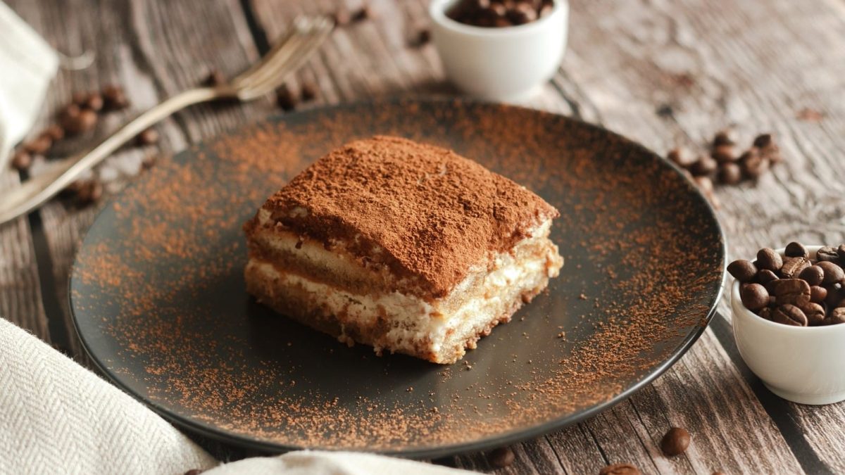 Ein Stück Tiramisu mit Baileys auf einem schwarzen Teller, mit Kakao-Pulver bestäubt, neben Schüsseln voller Kaffeebohnen