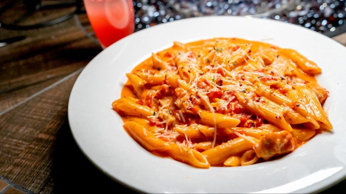 Italianische Pasta alla Vodka in einem weißen Teller auf einem dunklen Holztisch neben einem Cocktail und einer brennenden Feuerschale.