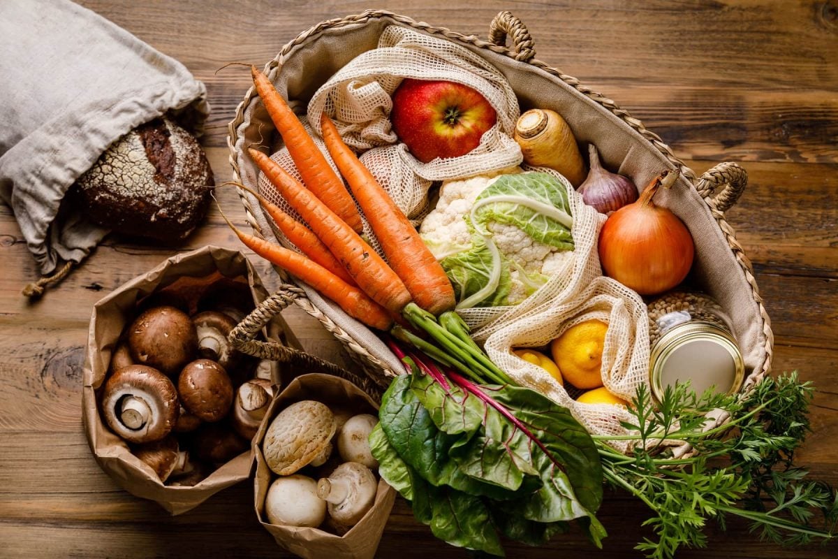Eine große Tasche, zwei Papiertüten und ein Beutel stehen auf einem dunkelbraunen Holztisch. Die Tasche ist gefüllt mit einer Variation aus Obst und Gemüse, darunter unter Anderem Karotten, Blumenkohl und Apfel. Die Papiertüten sind gefüllt mit Pilzen. In dem Beutel am linken Bildrand befindet sich ein Brot.